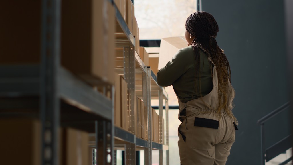 Chief logistics officer in warehouse arranging cardboard boxes full of merchandise ready to be shipped to clients. Meticulous director in depository using ladder to put parcels on cabinet shelves